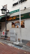 A street scene depicting a corner of a building with signage for 'Mogambo Singapore' and 'Canton St' visible. The exterior features street lamps and a no entry sign in the foreground. A billboard advertises food with images of dishes.