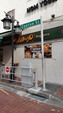 A street scene depicting a corner of a building with signage for 'Mogambo Singapore' and 'Canton St' visible. The exterior features street lamps and a no entry sign in the foreground. A billboard advertises food with images of dishes.