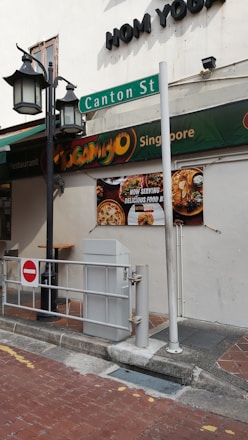 A street scene depicting a corner of a building with signage for 'Mogambo Singapore' and 'Canton St' visible. The exterior features street lamps and a no entry sign in the foreground. A billboard advertises food with images of dishes.