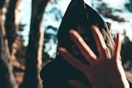 A mysterious figure wearing a dark hooded cloak stands in a forest, partially obscured by shadows. The person's face is painted in a sinister manner, with stark white eyes creating an eerie effect. A hand is raised in the foreground, partially covering the face, enhancing the enigmatic and slightly threatening atmosphere.