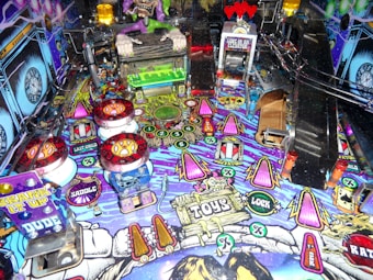 A vibrant pinball machine playfield featuring colorful graphics and electronic components, dominated by cartoonish monster figures and playful elements. The surface is densely packed with buttons, flashing lights, and ramps, creating an energetic and chaotic aesthetic.