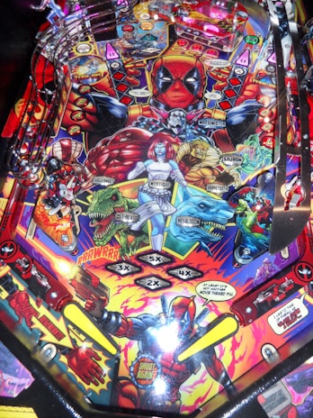 A colorful pinball machine playfield features a comic book theme with various superhero characters and villains, including a prominent red-masked figure at the top center. There are vibrant illustrations of characters like Juggernaut, Mystique, and others, surrounded by dynamic comic-style text and vibrant graphics. The playfield includes various ramps, targets, and lanes decorated with red, blue, yellow, and green shades.