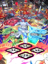 A colorful pinball machine playfield featuring comic-style artwork with various characters and creatures. There are detailed depictions of characters like Mystique and others, alongside creatures such as T-Rex and Megalodon. Numbered multipliers (2x, 3x, 4x, 5x) are prominently displayed as part of the game features.