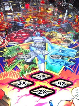 A colorful pinball machine playfield featuring comic-style artwork with various characters and creatures. There are detailed depictions of characters like Mystique and others, alongside creatures such as T-Rex and Megalodon. Numbered multipliers (2x, 3x, 4x, 5x) are prominently displayed as part of the game features.