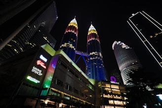 A night scene featuring the illuminated Petronas Twin Towers with surrounding skyscrapers. The towers display vibrant lights, and there are signs of various brands like Marks and Spencer.