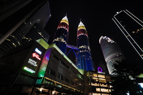 A night scene featuring the illuminated Petronas Twin Towers with surrounding skyscrapers. The towers display vibrant lights, and there are signs of various brands like Marks and Spencer.