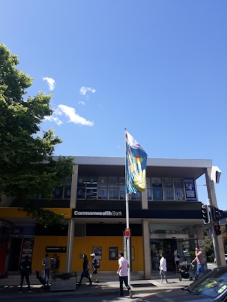 A modern building with a Commonwealth Bank sign is visible, accompanied by a bustling street with several pedestrians walking by. The sky is clear with a few clouds, and a large colorful flag is mounted on a pole in front of the building. Tree foliage is visible on the left side of the image.
