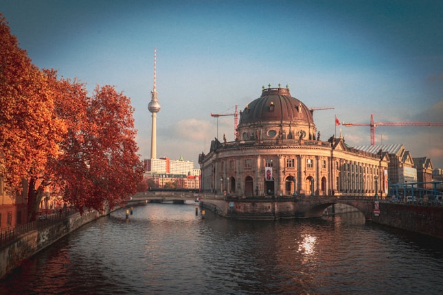 A large, historic museum building is situated by a tranquil river. The architecture features a prominent dome and classical design elements. In the background, a tall television tower rises against a clear blue sky. The scene is accented with the warm hues of autumn foliage on nearby trees, and several construction cranes can be seen, indicating ongoing development in the area.
