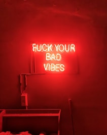 A neon sign with bold red letters displaying the phrase 'FUCK YOUR BAD VIBES' is mounted on a wall. The intense red glow of the sign illuminates the surrounding area, creating a vibrant and edgy atmosphere. The electrical wiring is visible and leads to the left of the frame. Below the sign is a shelf with some objects that are not clearly discernible.