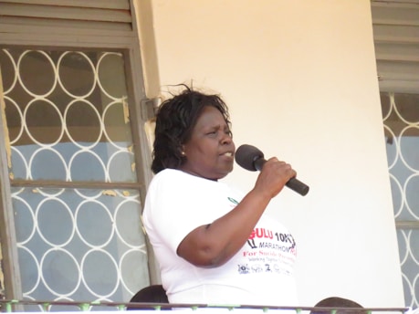 A person is speaking into a microphone, standing on a balcony or platform next to a window with decorative metalwork. They are wearing a white shirt with text.