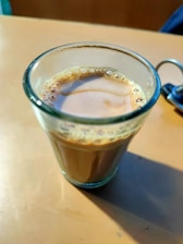 A glass cup filled with chai is placed on a light brown surface. The tea appears frothy at the top, and there's a metal keyring partially visible on the right side.