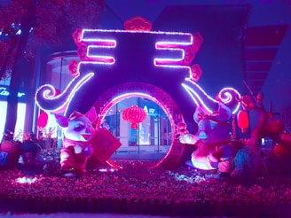 A vibrant and colorful nighttime display featuring whimsical pig figures adorned in festive attire, surrounded by illuminated decorations and traditional red lanterns. The centerpiece is an arch-like structure with intricate designs, beautifully lit with neon purple and pink lights, creating a lively and celebratory atmosphere.