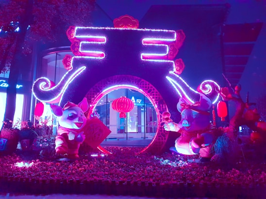 A vibrant and colorful nighttime display featuring whimsical pig figures adorned in festive attire, surrounded by illuminated decorations and traditional red lanterns. The centerpiece is an arch-like structure with intricate designs, beautifully lit with neon purple and pink lights, creating a lively and celebratory atmosphere.