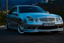 A silver luxury sedan is parked on a dark road with mountainous terrain in the background. The car has a sleek design with prominent front grilles and alloy wheels. The lighting suggests early evening, casting shadows across the vehicle.