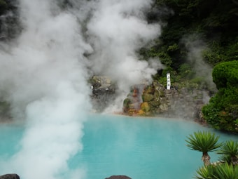 A geothermal hot spring pool emits thick steam against a backdrop of lush greenery and rock formations. The water appears bright blue, and the dense vapor creates a mystical atmosphere. A sign with text is visible near the rocky area.