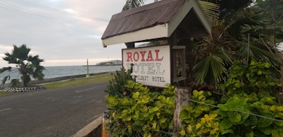 A sign reading 'ROYAL HOTEL, Your Budget Hotel' is surrounded by lush green foliage. The sign is positioned next to a coastal road, with the ocean visible in the background. Palm trees are present, and the scene is set against a cloudy sky.