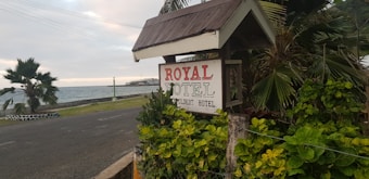 A sign reading 'ROYAL HOTEL, Your Budget Hotel' is surrounded by lush green foliage. The sign is positioned next to a coastal road, with the ocean visible in the background. Palm trees are present, and the scene is set against a cloudy sky.