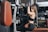 A woman is exercising in a gym setting, using a cable machine. She is seated on a bench and is pulling down on a handle attached to the machine, wearing athletic gear including a black sports bra and leggings.