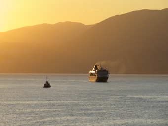 A large cruise ship and a smaller boat are sailing on calm waters during sunset. The horizon is dominated by rolling hills creating a serene backdrop. The sky is filled with warm hues of orange and yellow, reflecting on the surface of the water.