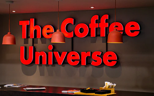 A bold, red 3D sign with the words 'The Coffee Universe' is mounted on a dark wall. Hanging from the ceiling are three orange pendant lamps. Below the sign, there is a dark brown counter with a stack of magazines and a small wooden container holding various items, such as packets and stirrers.