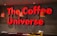 A bold, red 3D sign with the words 'The Coffee Universe' is mounted on a dark wall. Hanging from the ceiling are three orange pendant lamps. Below the sign, there is a dark brown counter with a stack of magazines and a small wooden container holding various items, such as packets and stirrers.