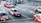 Several red taxis with white tops are driving on a multi-lane road. The road is marked with arrows indicating different lane directions. Other vehicles are visible, including a dark-colored van. The street features a sidewalk with a metal railing and a pedestrian. Buildings and urban infrastructure are in the background.