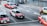 Several red taxis with white tops are driving on a multi-lane road. The road is marked with arrows indicating different lane directions. Other vehicles are visible, including a dark-colored van. The street features a sidewalk with a metal railing and a pedestrian. Buildings and urban infrastructure are in the background.