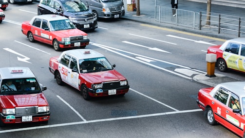 Several red taxis with white tops are driving on a multi-lane road. The road is marked with arrows indicating different lane directions. Other vehicles are visible, including a dark-colored van. The street features a sidewalk with a metal railing and a pedestrian. Buildings and urban infrastructure are in the background.