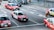 Several red taxis with white tops are driving on a multi-lane road. The road is marked with arrows indicating different lane directions. Other vehicles are visible, including a dark-colored van. The street features a sidewalk with a metal railing and a pedestrian. Buildings and urban infrastructure are in the background.