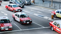 Several red taxis with white tops are driving on a multi-lane road. The road is marked with arrows indicating different lane directions. Other vehicles are visible, including a dark-colored van. The street features a sidewalk with a metal railing and a pedestrian. Buildings and urban infrastructure are in the background.