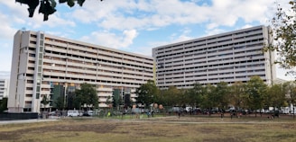 A large residential building with multiple floors and balconies is seen, surrounded by green trees and an open grassy area. People can be observed engaging in outdoor activities such as playing and walking in the foreground. The sky is partly cloudy, providing a serene backdrop to the urban scene.