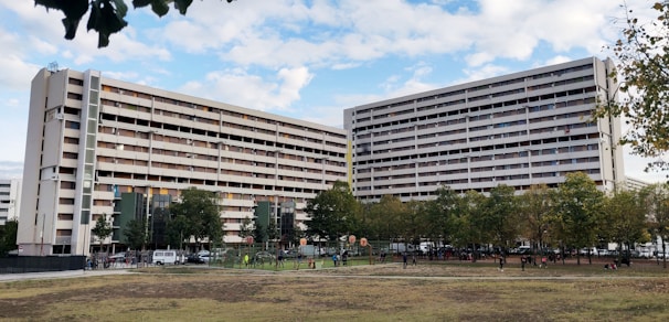 A large residential building with multiple floors and balconies is seen, surrounded by green trees and an open grassy area. People can be observed engaging in outdoor activities such as playing and walking in the foreground. The sky is partly cloudy, providing a serene backdrop to the urban scene.