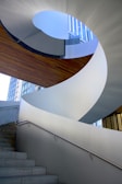 A modern architectural spiral staircase with sleek white surfaces and a metal handrail. The stairs lead upward through an open space, with natural light shining through a large window in the background. The design is both functional and artistic, emphasizing curves and clean lines.