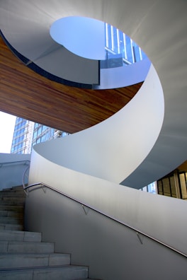 A modern architectural spiral staircase with sleek white surfaces and a metal handrail. The stairs lead upward through an open space, with natural light shining through a large window in the background. The design is both functional and artistic, emphasizing curves and clean lines.