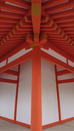 A detailed architectural view of a structure with vibrant orange beams and white panels, forming a symmetrical and geometric pattern. The beams converge at the ceiling, creating a striking visual impact with clean lines and a modern design.