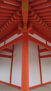 A detailed architectural view of a structure with vibrant orange beams and white panels, forming a symmetrical and geometric pattern. The beams converge at the ceiling, creating a striking visual impact with clean lines and a modern design.