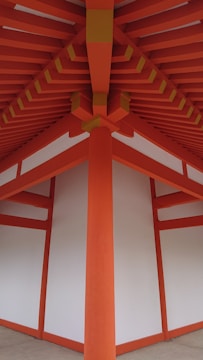 A detailed architectural view of a structure with vibrant orange beams and white panels, forming a symmetrical and geometric pattern. The beams converge at the ceiling, creating a striking visual impact with clean lines and a modern design.