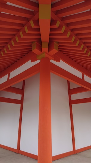 A detailed architectural view of a structure with vibrant orange beams and white panels, forming a symmetrical and geometric pattern. The beams converge at the ceiling, creating a striking visual impact with clean lines and a modern design.