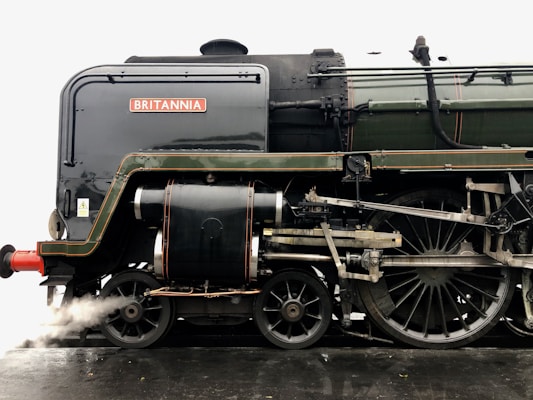 A close-up view of a vintage steam locomotive with intricate mechanical details. The metal surface is dark, with various components and pipes visible. The word 'BRITANNIA' is displayed prominently on the side.