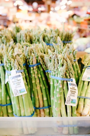 Bundles of fresh asparagus tied with blue rubber bands are displayed in a supermarket setting. Price tags are attached to some of the bundles, and the background shows a blurred interior of the store with warm lighting.