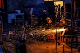 A worker wearing protective gear is engaged in metalwork, creating bright orange sparks as material is cut or welded. The scene is set in a dimly lit industrial space with various tanks and metal structures scattered around. The background includes a partially open door with warm yellow light illuminating the interior.