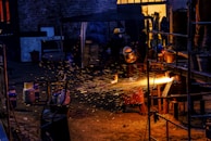 A worker wearing protective gear is engaged in metalwork, creating bright orange sparks as material is cut or welded. The scene is set in a dimly lit industrial space with various tanks and metal structures scattered around. The background includes a partially open door with warm yellow light illuminating the interior.