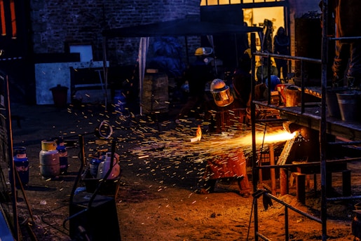 A worker wearing protective gear is engaged in metalwork, creating bright orange sparks as material is cut or welded. The scene is set in a dimly lit industrial space with various tanks and metal structures scattered around. The background includes a partially open door with warm yellow light illuminating the interior.