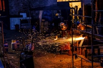 A worker wearing protective gear is engaged in metalwork, creating bright orange sparks as material is cut or welded. The scene is set in a dimly lit industrial space with various tanks and metal structures scattered around. The background includes a partially open door with warm yellow light illuminating the interior.