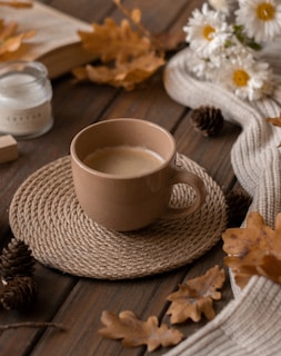 A warm cup of coffee sits atop a woven coaster on a wooden table surrounded by autumn-themed decor, including dried leaves, pinecones, and white daisies. A cozy knitted scarf is draped nearby, along with a lit candle and an open book, creating a rustic and inviting atmosphere.