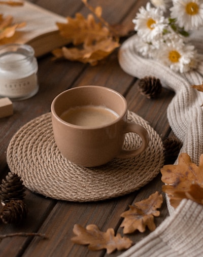 A warm cup of coffee sits atop a woven coaster on a wooden table surrounded by autumn-themed decor, including dried leaves, pinecones, and white daisies. A cozy knitted scarf is draped nearby, along with a lit candle and an open book, creating a rustic and inviting atmosphere.