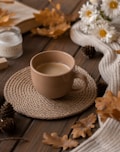 A warm cup of coffee sits atop a woven coaster on a wooden table surrounded by autumn-themed decor, including dried leaves, pinecones, and white daisies. A cozy knitted scarf is draped nearby, along with a lit candle and an open book, creating a rustic and inviting atmosphere.
