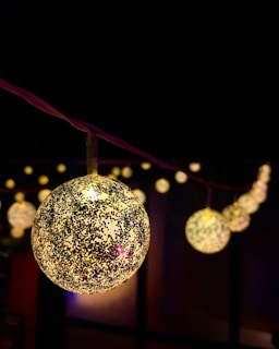 A close-up view of decorative spherical lights adorned with a speckled pattern, set against a dark background. The lights are strung along a purple cord, creating a festive and cozy atmosphere with blurred lights in the background.