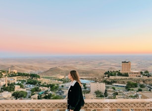 A person stands on a balcony overlooking a vast landscape with a panoramic view of rolling hills and a small town below. The sky displays a gradient of pastel colors as the sun sets, creating a serene and peaceful ambiance. Buildings surrounded by greenery are visible, with one prominent structure in the distance.