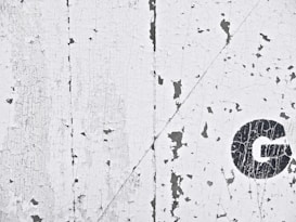The image depicts a textured surface with white, peeling paint revealing an underlying dark layer. On the right side, a black letter 'G' is partially visible.